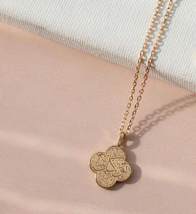 Gold necklace with a clover-shaped pendant with arabic engraving on a light pink surface