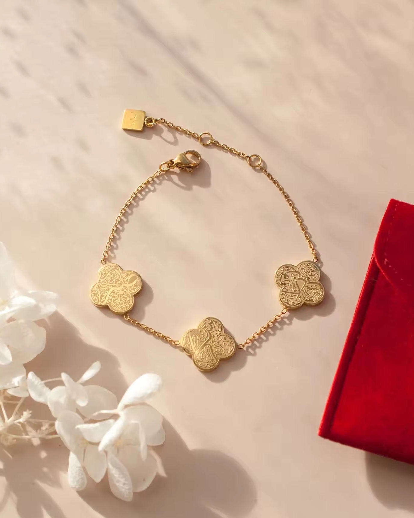 Gold bracelet with clover design and arabic text on a beige surface with white flowers and a red box