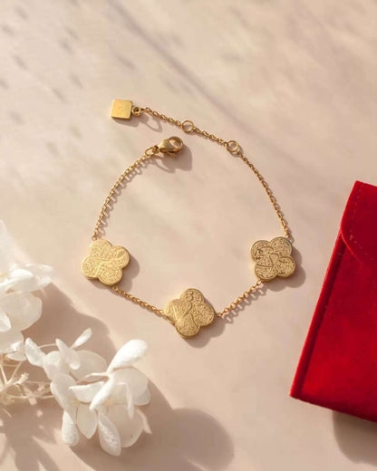 Gold bracelet with clover design and arabic text on a beige surface with white flowers and a red box