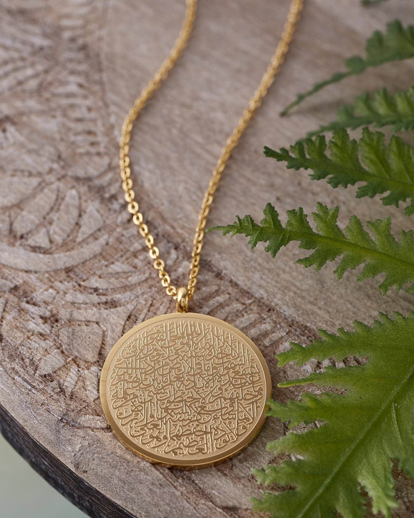 Gold necklace with a round pendant on a wooden surface with a fern leaf