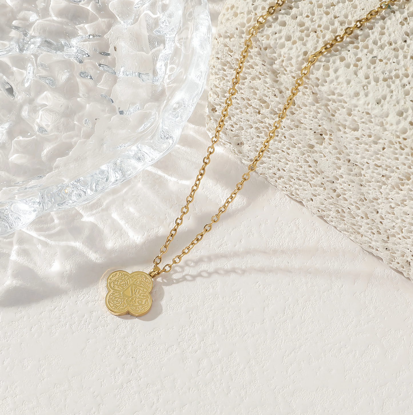 Gold necklace with a clover-shaped pendant with arabic engraving  on a textured white surface